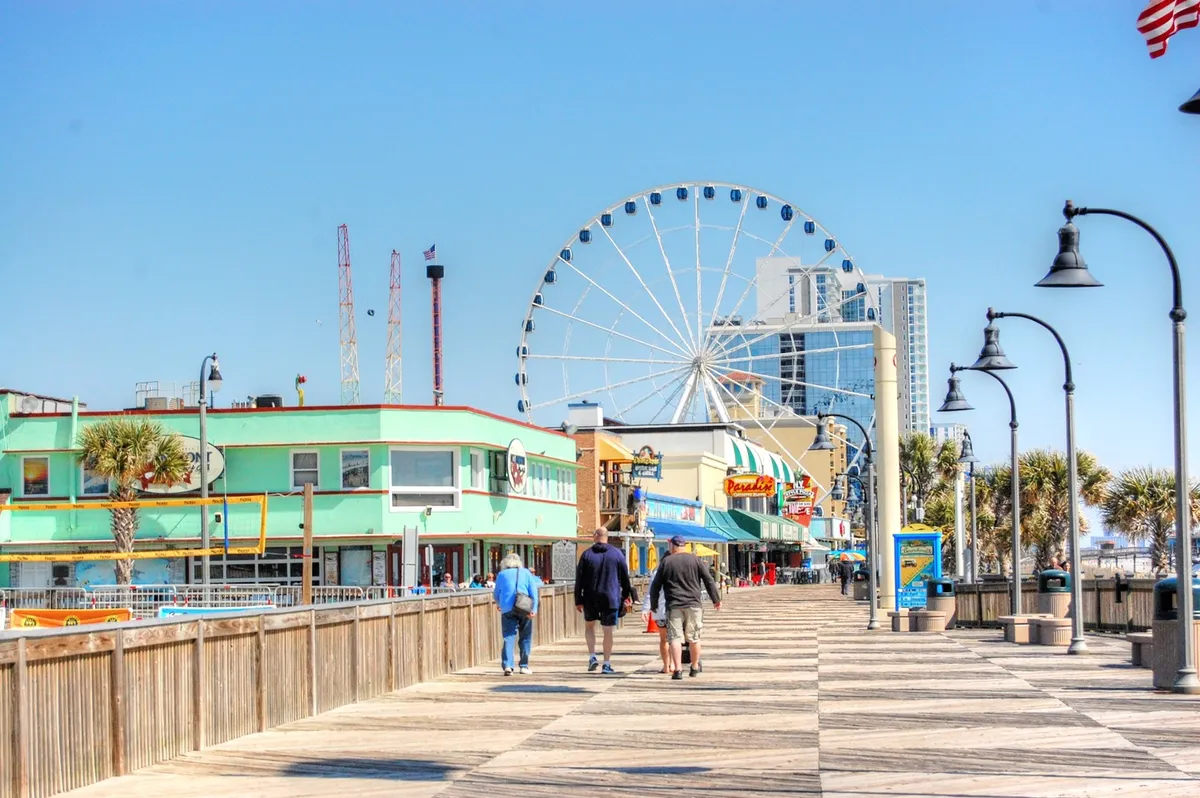 A lively shot of the Myrtle Beach boardwalk, featuring a brightly lit ferris wheel, colorful storefronts, and vacationers strolling along the pier. The image exudes energy and fun, with people enjoying classic seaside attractions and the festive atmosphere.