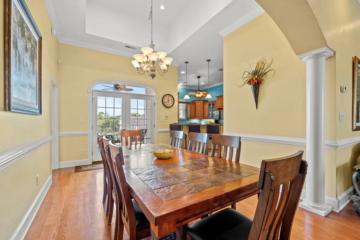 A bright, cheerful dining area with a large table set beneath a sparkling chandelier. Large windows allow natural light to fill the room, while the adjacent kitchen area and wall decorations add to the open, spacious feel. This is a space designed for family dinners, celebrations, and shared moments.