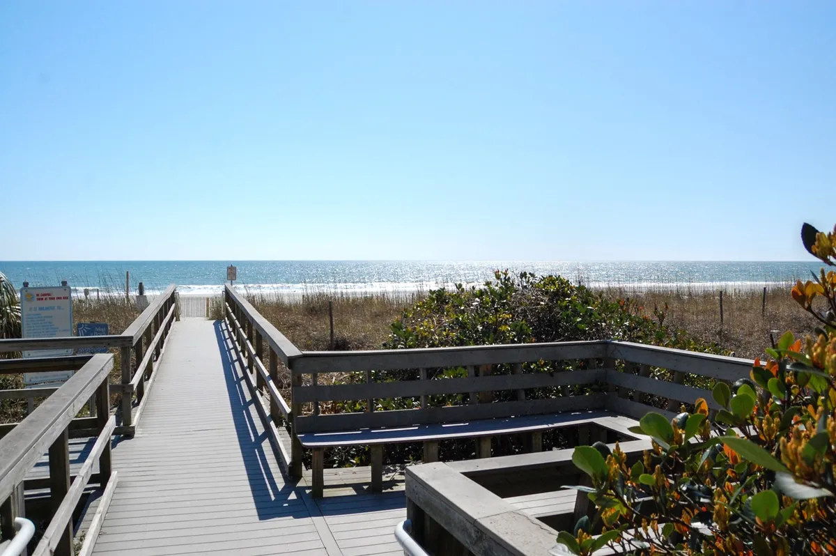 A picturesque wooden walkway winds through dune grass, leading directly to a wide, open view of the ocean under a clear blue sky. The scene is serene and beckons exploration, symbolizing both the literal and figurative journey from home to the beach.