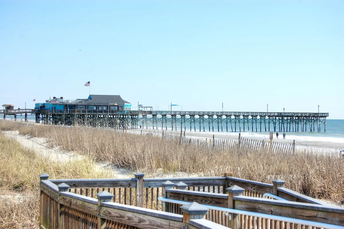 A picturesque pier stretches over the beach toward the ocean, with a flag waving and grasses in the foreground. The beach is calm and inviting, perfect for walking, fishing, or simply soaking up the atmosphere.