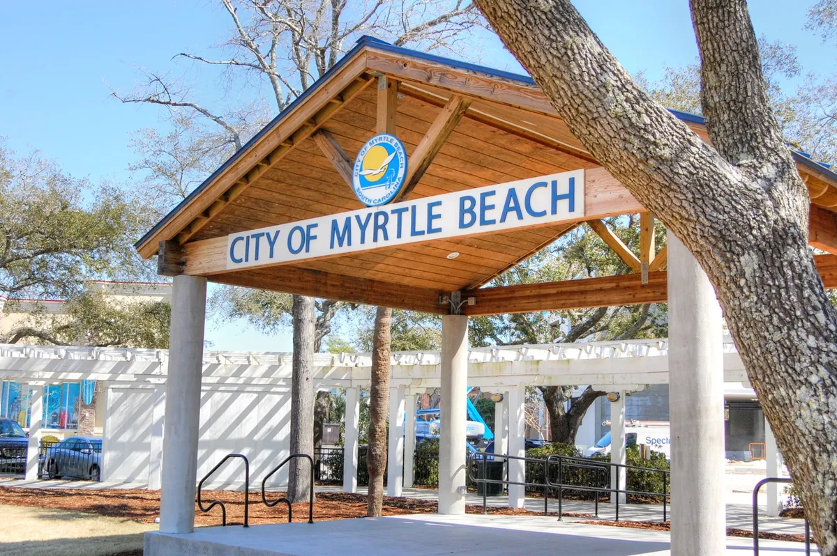 This image captures the inviting 'City of Myrtle Beach' sign situated under a wooden structure, surrounded by lush trees. The scene suggests a sense of arrival and local community, with the possibility of tourism and public gatherings.