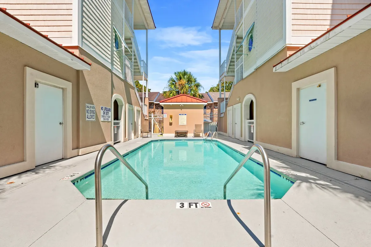 A sparkling outdoor community pool framed by lounge chairs and surrounded by resort-style buildings. The scene captures guests swimming, sunbathing, and gathering poolside, evoking a relaxed and social atmosphere.