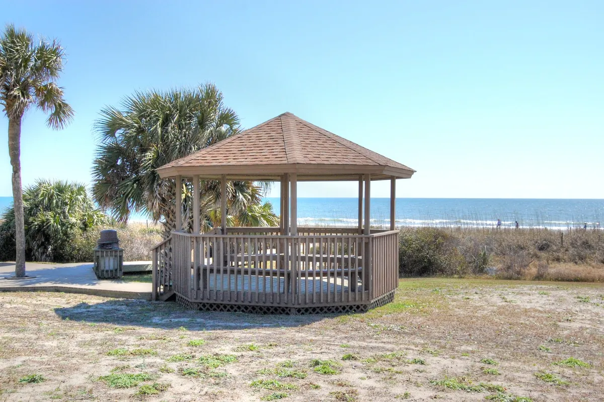 A scenic wooden gazebo overlooks a sparkling ocean framed by palm trees, evoking relaxation and the coastal lifestyle. The space feels ideal for social gatherings or casual strolls with friends.