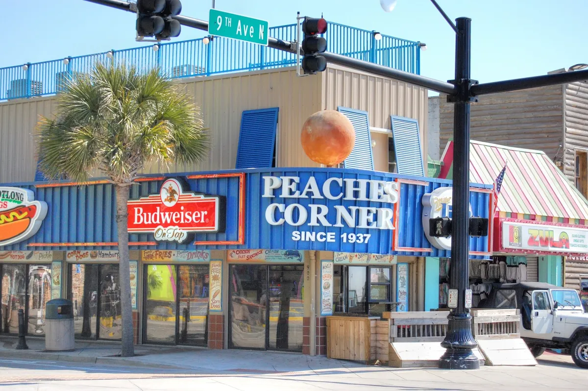A lively streetscape highlights the colorful facades of Myrtle Beach’s legendary Peaches Corner, flanked by palm trees and neon signage. People gather outdoors, enjoying food, drinks, and the energetic atmosphere. This image captures the festive spirit and abundance of dining and nightlife options just a walk away.