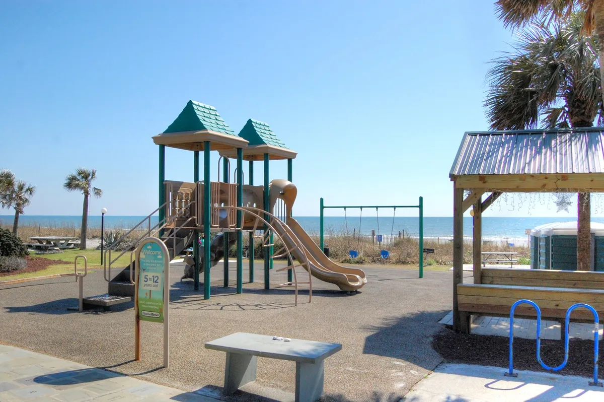 A cheerful playground scene overlooking the ocean and framed by palm trees, with kids playing on swings and equipment while families gather nearby. The beachside setting emphasizes relaxation and outdoor fun for children and parents alike.