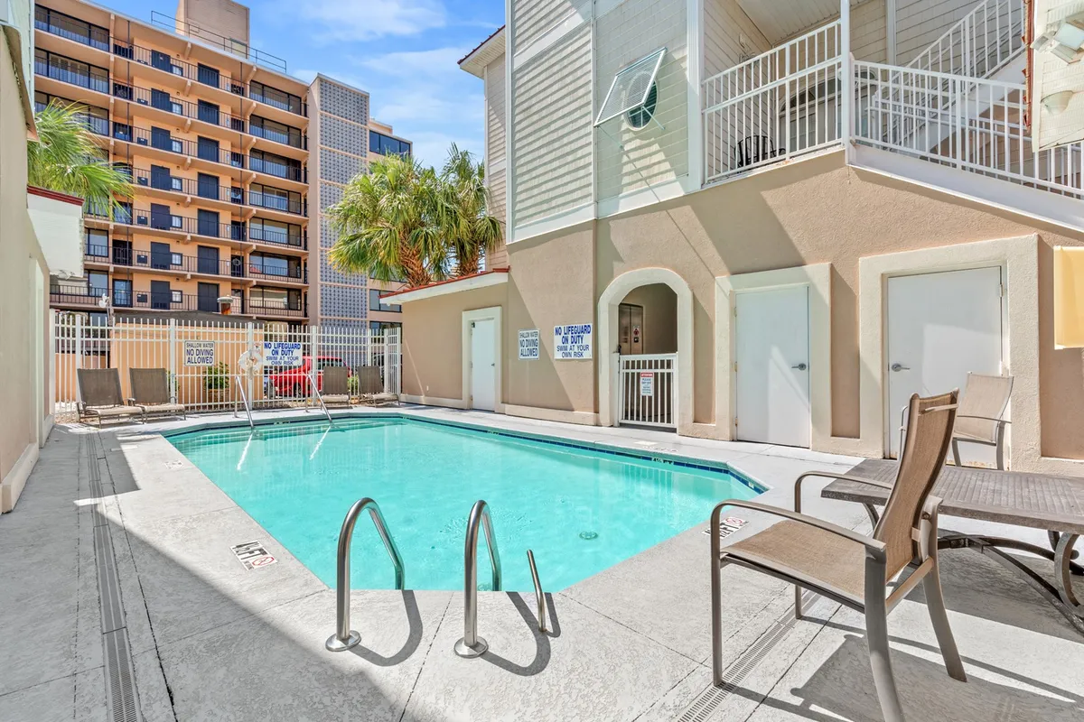A bright, inviting view of the community swimming pool with plenty of poolside chairs, palm trees, and a clear look at the building facade. The setting is lively, with guests enjoying the water and mingling by the pool.