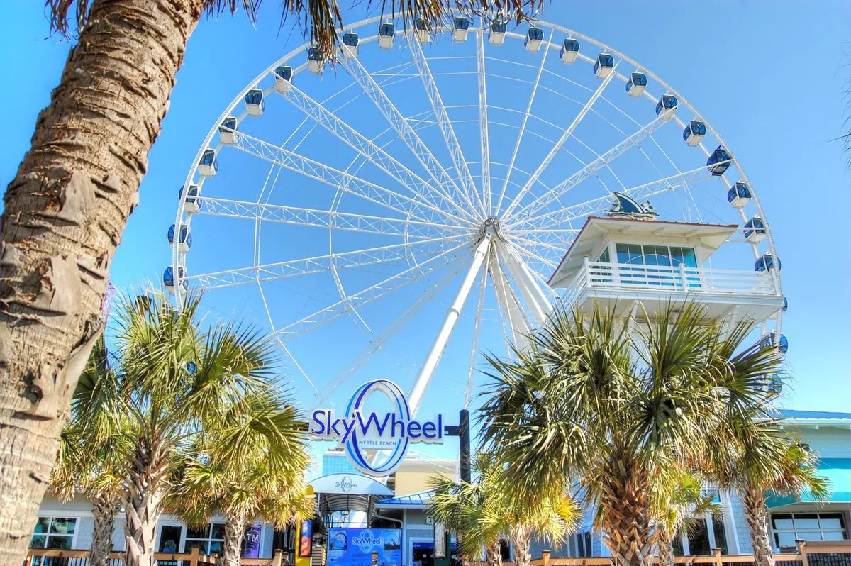 The iconic SkyWheel rises above palm trees against a bright blue sky—a lively landmark that promises fun and panoramic views. The scene is vibrant and energetic, hinting at the excitement that awaits just beyond the beach.