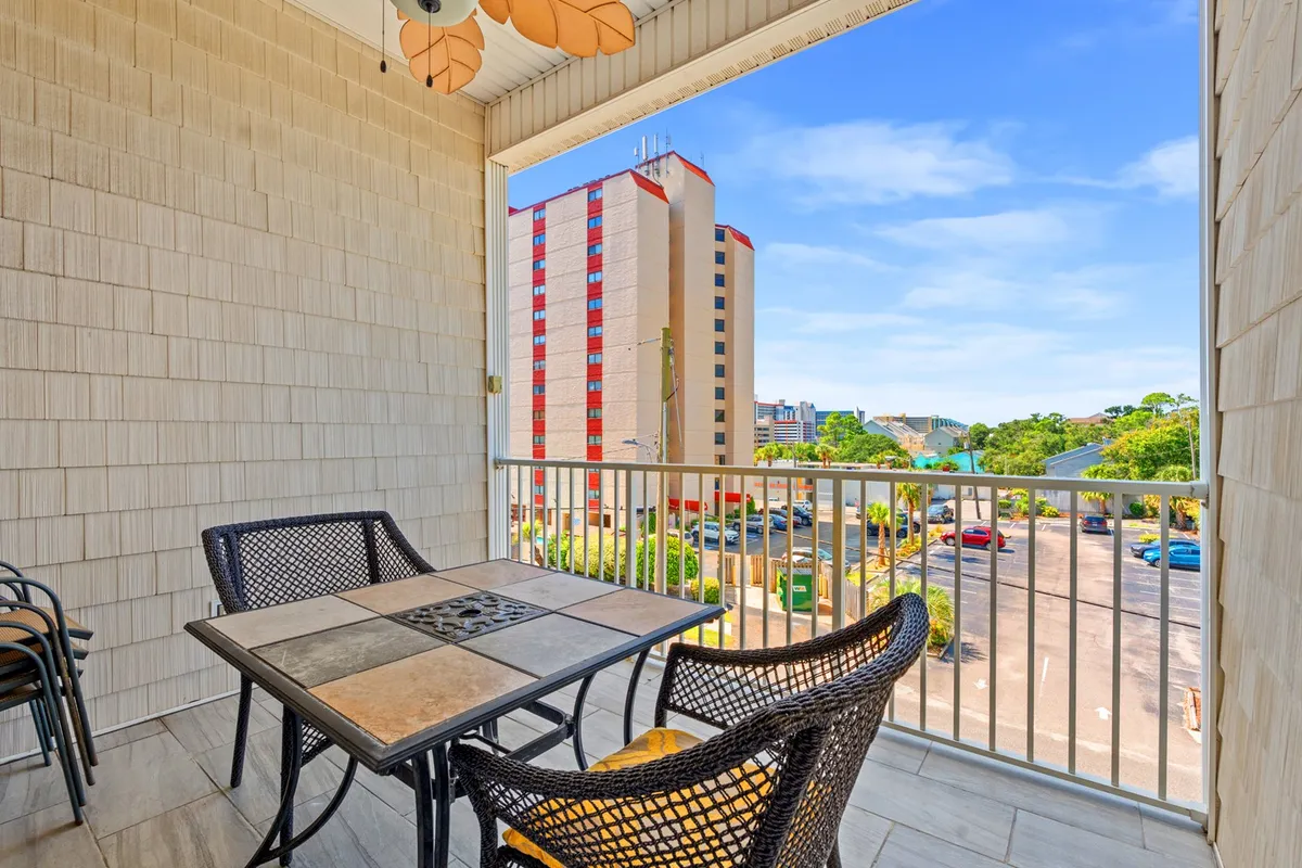An inviting outdoor dining setup features a table with comfortable chairs, accented by a decorative ceiling fan overhead. The scene opens to a city view, blending the private comfort of home with the vibrant energy of Myrtle Beach just beyond. The warm, ambient lighting suggests an ideal spot for evening gatherings or relaxing after a day of exploration.