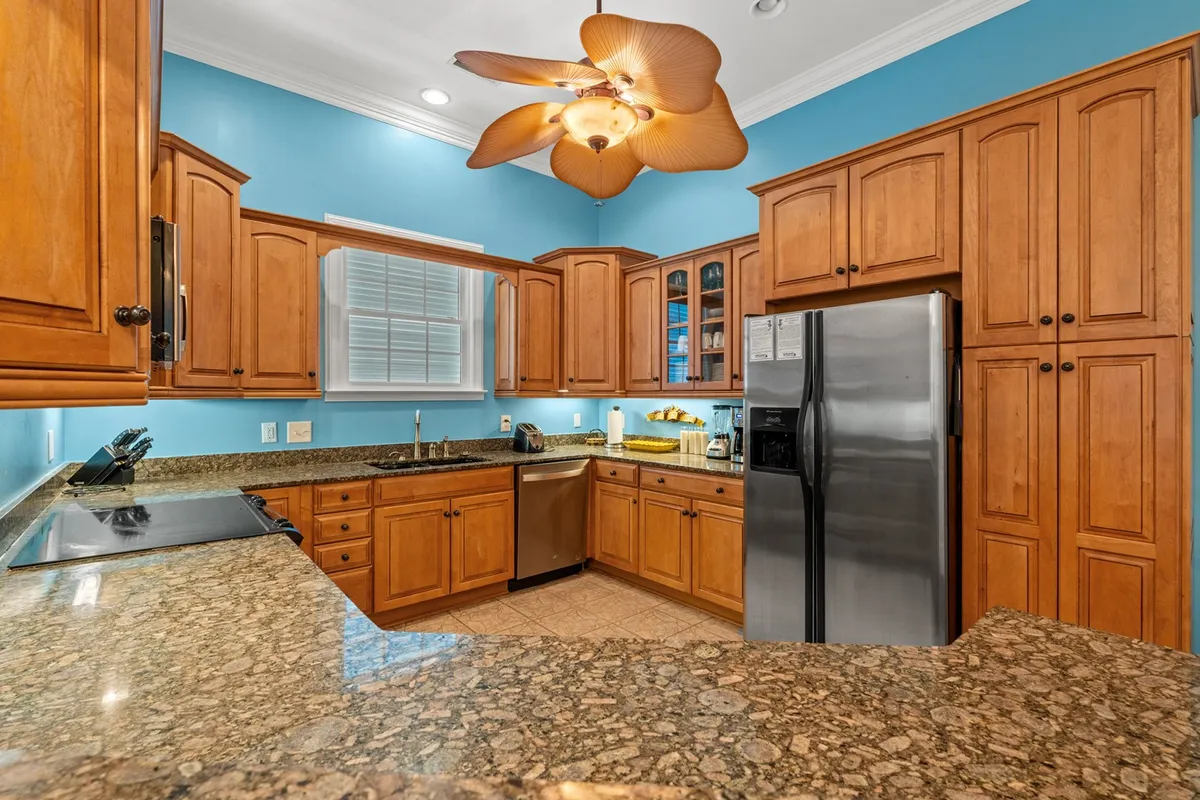 A sleek kitchen with granite countertops, wooden cabinetry, and island seating. A ceiling fan overhead adds comfort, and the island provides a perfect spot for casual snacks or conversation. The kitchen is spacious and modern, seamlessly connecting to the rest of the living area.